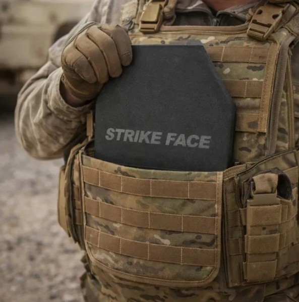 A gloved hand inserts a black armor plate into a camouflage plate carrier