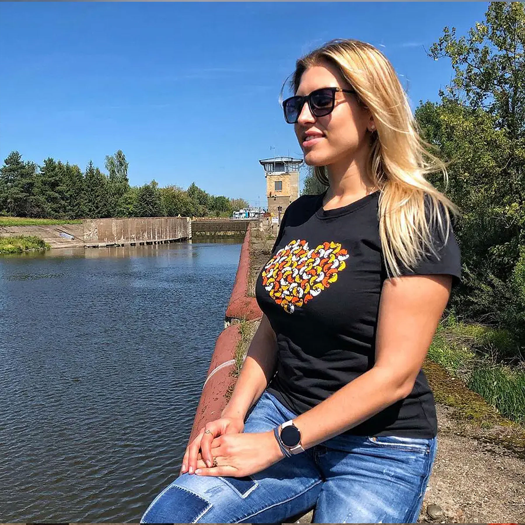 A woman wearing a black T-shirt with a multicolored heart on the chest is sitting on the riverbank