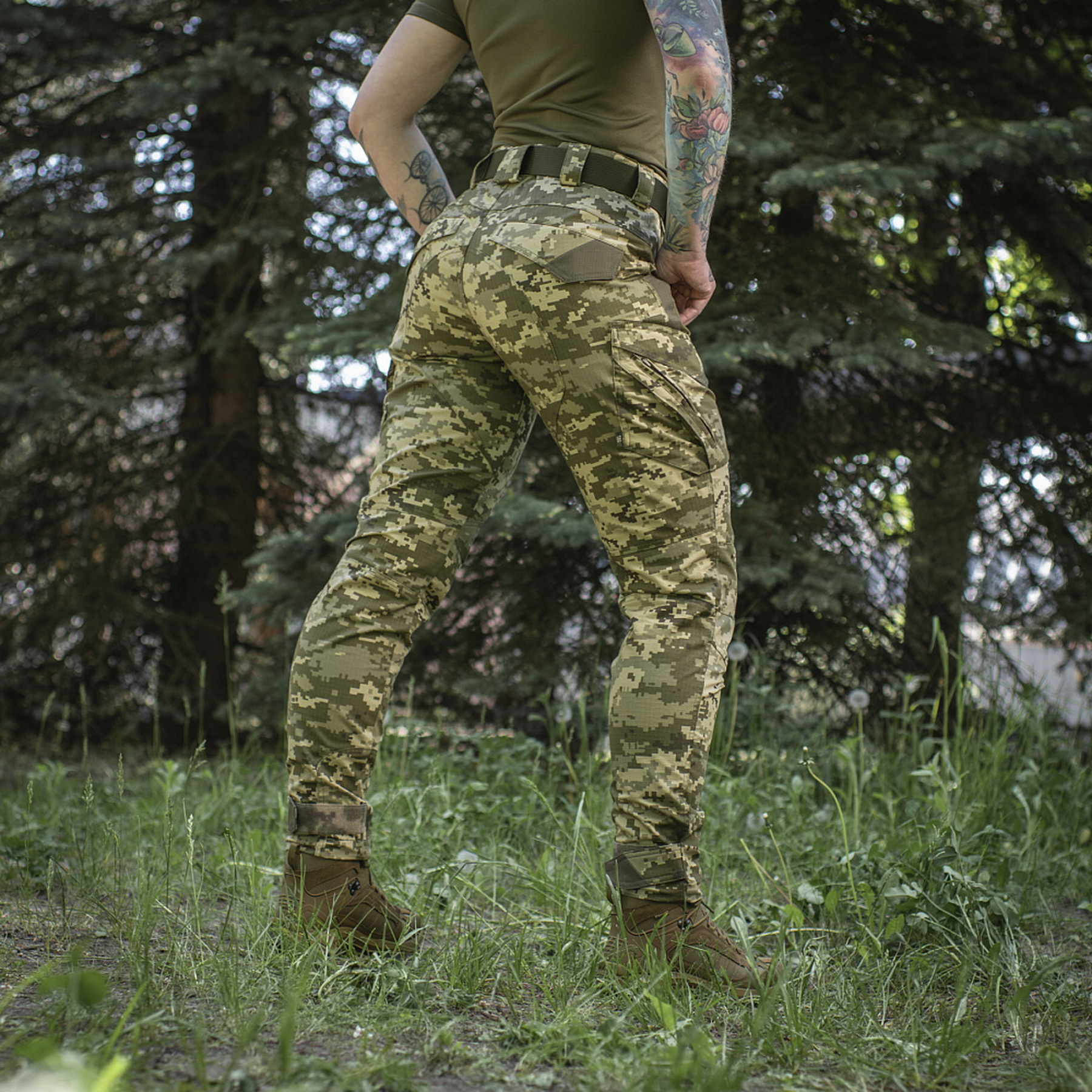 A woman wearing pixel-patterned tactical pants against a backdrop of trees (view from behind)
