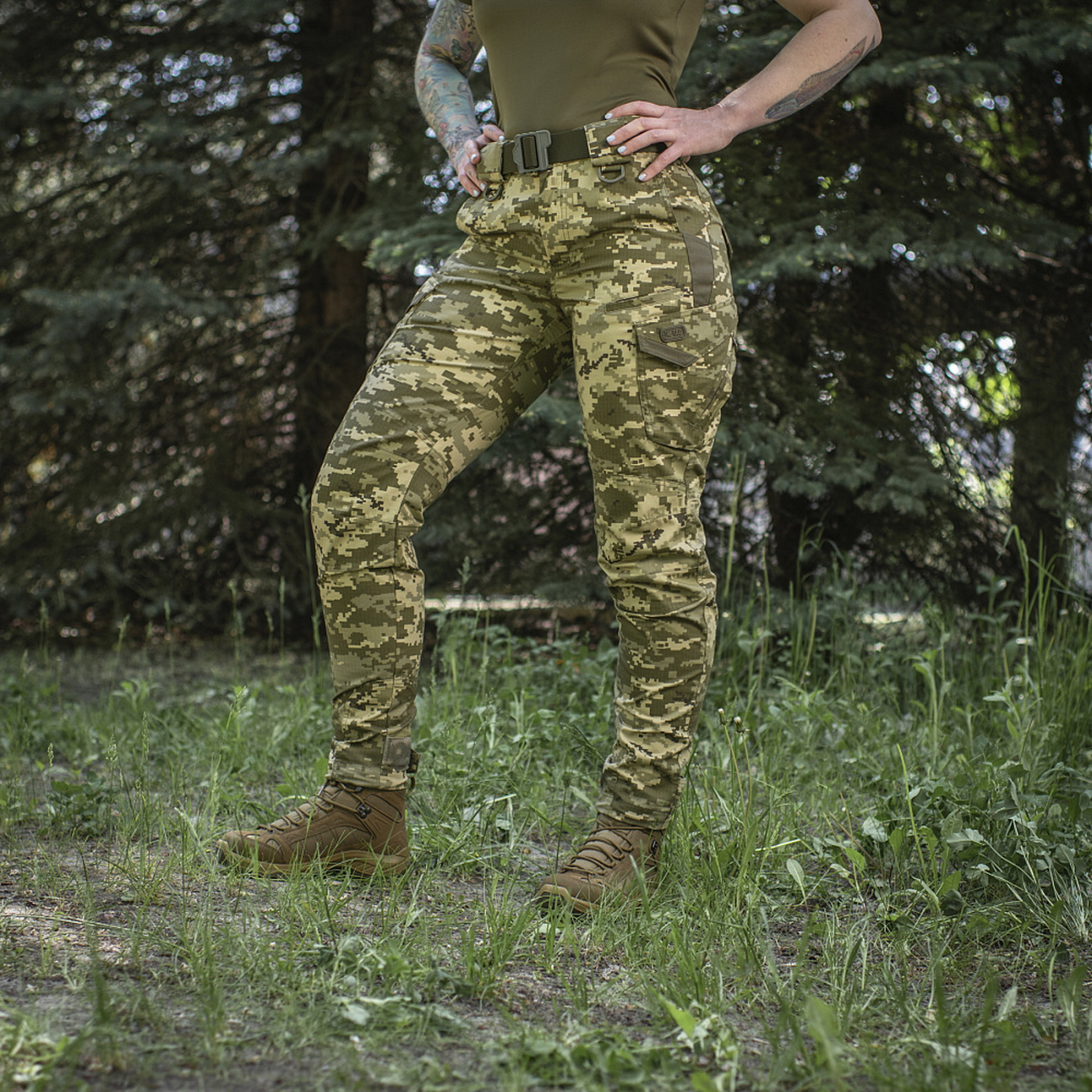 A woman wearing pixel-patterned tactical pants against a backdrop of trees (front view)