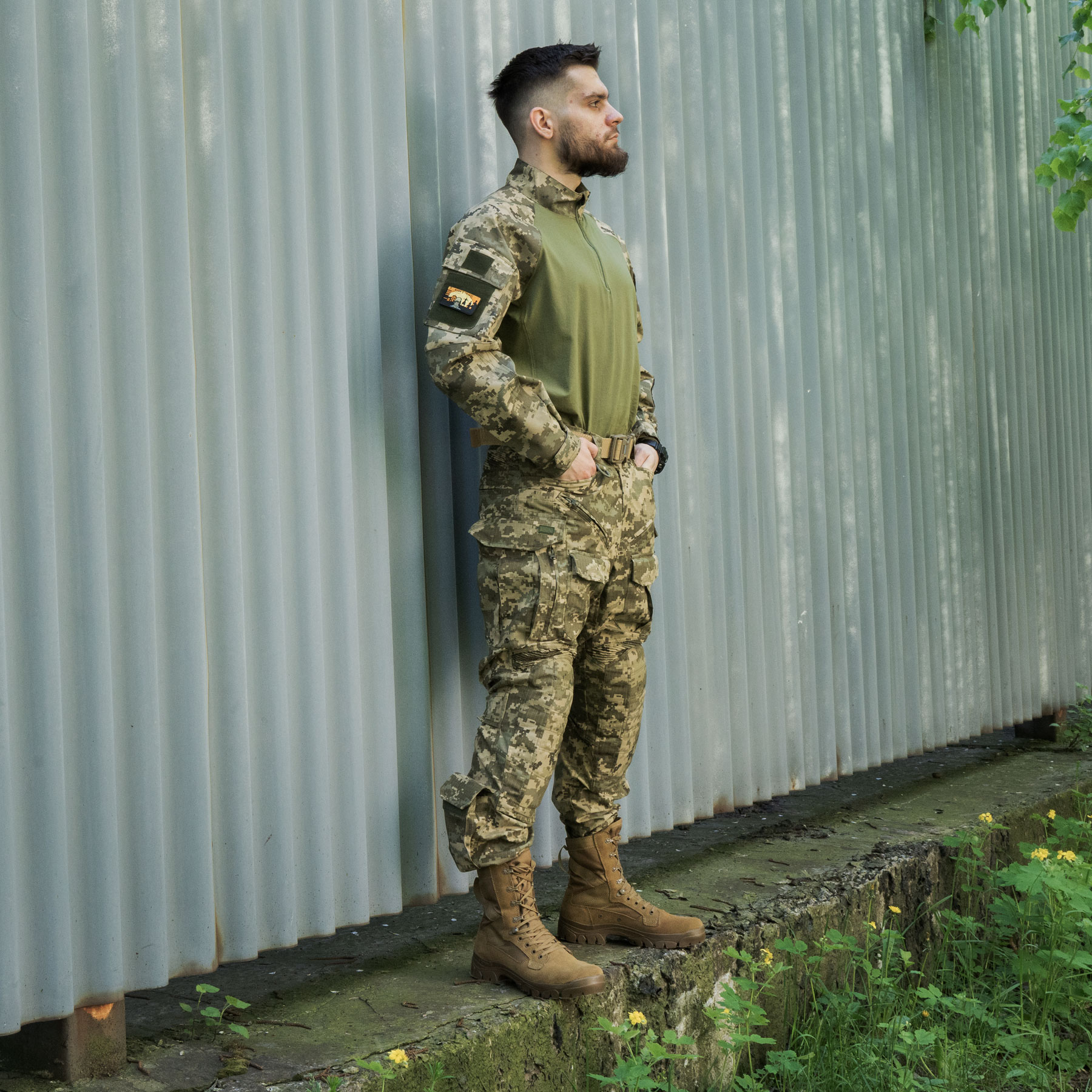 A man wearing a tactical shirt and pixel-patterned pants is standing near a corrugated wall