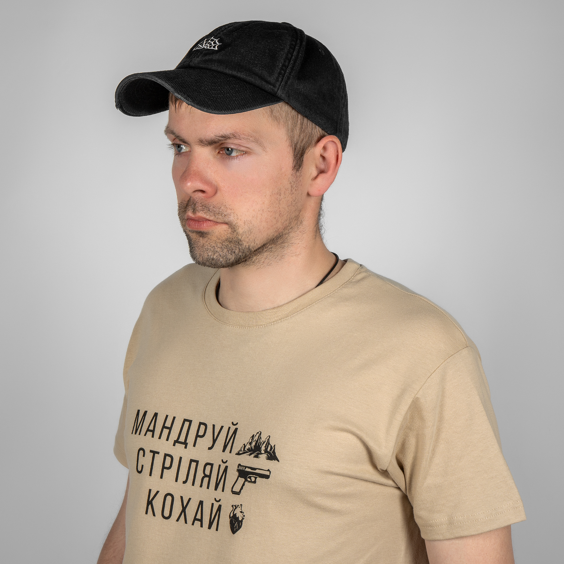 Мужчина в черной кепке с белой паутиной бренда и футболке "Путешествуй. Стреляй. Люби."