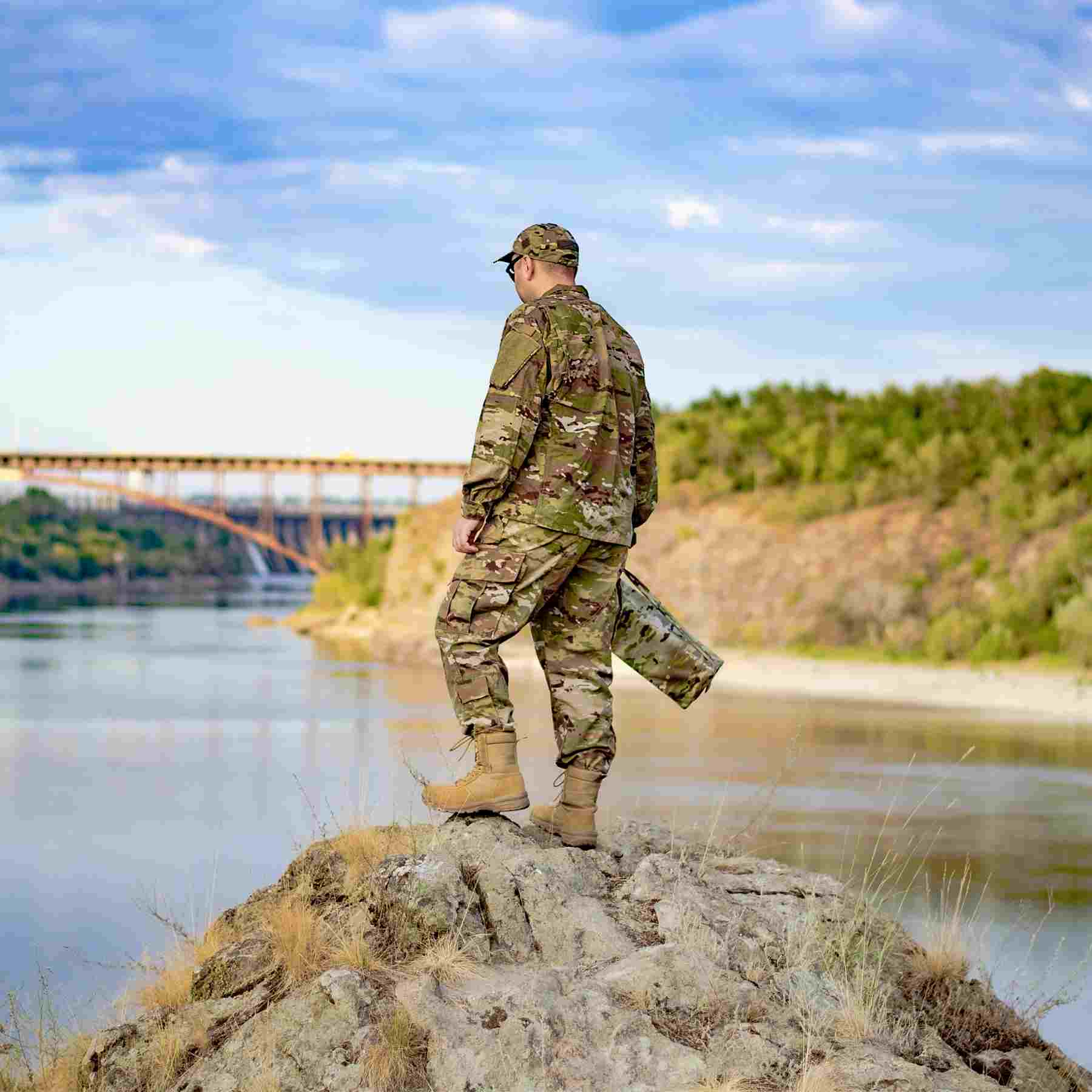 A person in military clothing stands on a rocky outcrop, looking at a bridge over the river