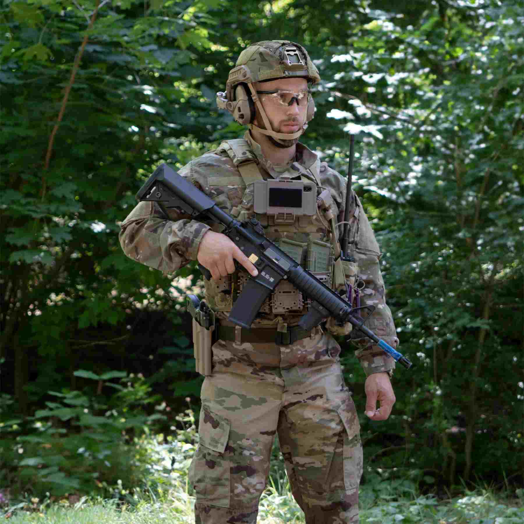 A soldier in military clothing and gear, wearing a helmet and protective glasses, holds a rifle in the forest